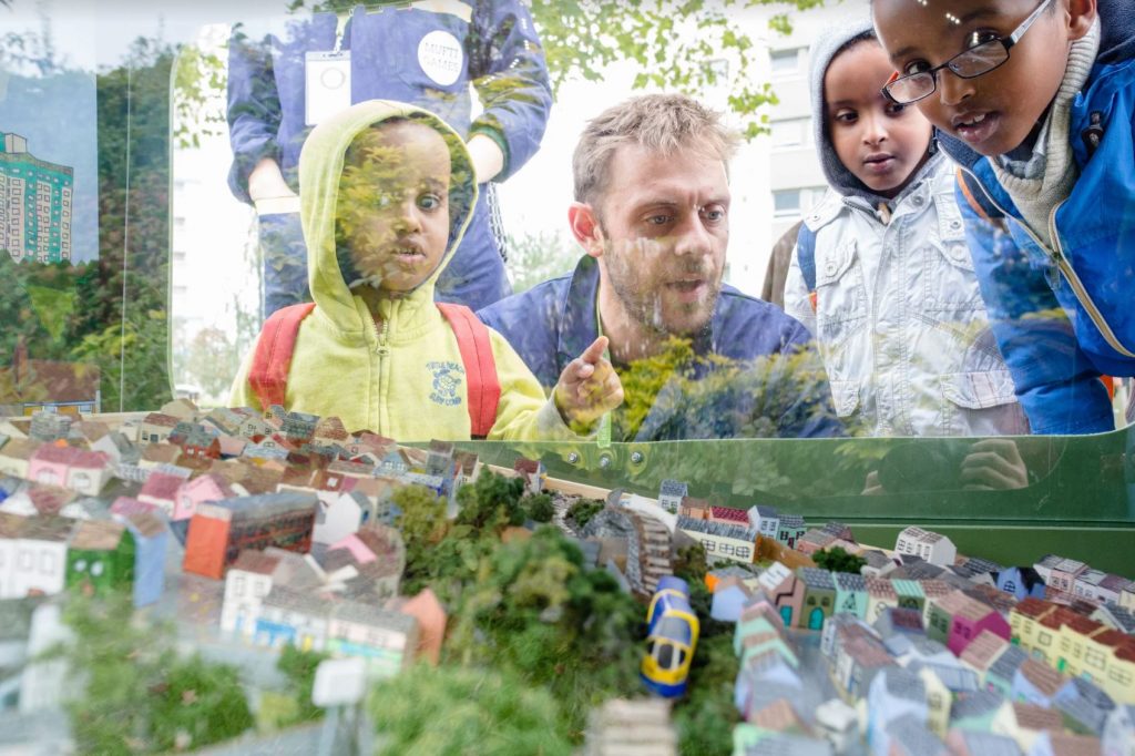 Adults and children look at a miniature model of Easton in Bristol. The model in behind plastic inside a large bin. The model is a marble run